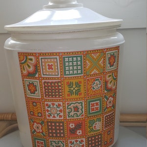 May include: A white ceramic storage jar with a lid, decorated with a patchwork design of floral and geometric patterns in orange, yellow, green, and brown. The jar has a rounded shape and a white knob.
