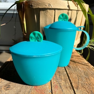 May include: Two turquoise plastic containers with lids. The containers have a green swirl design on the lids. The containers are sitting on a wooden surface.