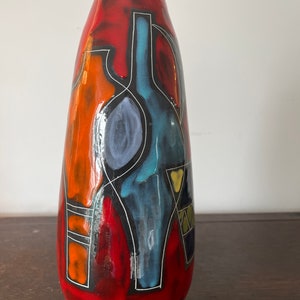 May include: A red ceramic vase with abstract geometric designs in blue, orange, and white. The vase has a rounded, elongated shape.