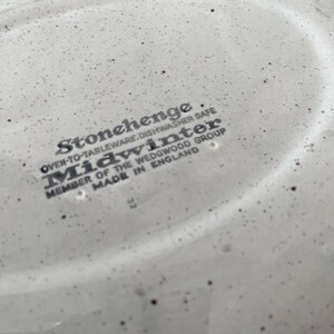 May include: Close-up of the bottom of a white ceramic plate with brown speckles. The plate has the text "Stonehenge Oven-to-Tableware, Dishwasher Safe Midwinter Member of the Wedgwood Group Made in England" printed on it.