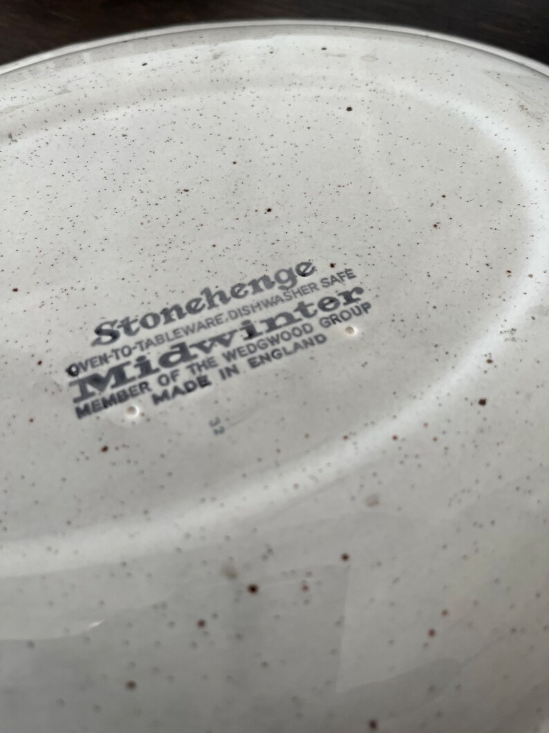 May include: Close-up of the bottom of a white ceramic plate with brown speckles. The plate has the text "Stonehenge Oven-to-Tableware, Dishwasher Safe Midwinter Member of the Wedgwood Group Made in England" printed on it.