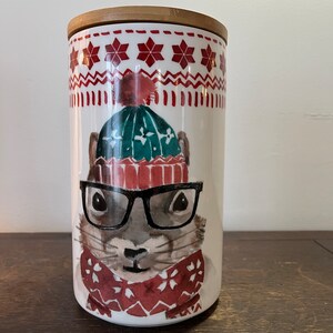 Peut inclure: Un pot en céramique blanc avec un couvercle en bambou. Le pot présente une illustration à l'aquarelle d'un écureuil portant un bonnet vert, des lunettes noires et une écharpe rouge. Le haut a un motif de flocons de neige rouges et blancs.
