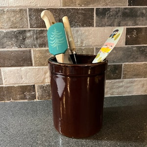 May include: A brown ceramic utensil holder with a wooden spatula, a green spatula with a pine tree design, and a colorful spatula with a design of a camper van.
