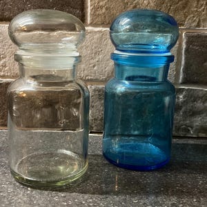 May include: Two glass jars with lids. The jar on the left is clear glass with a white lid. The jar on the right is blue glass with a blue lid.