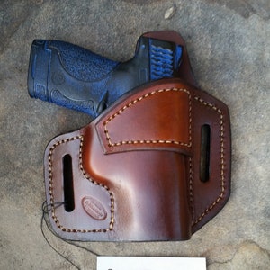 May include: A brown leather holster with a blue and black handgun inside. The holster is stitched with light-colored thread and has a tag that reads "CUSTOM MADE BY Self Deo Gloria JOHN JACKSON."
