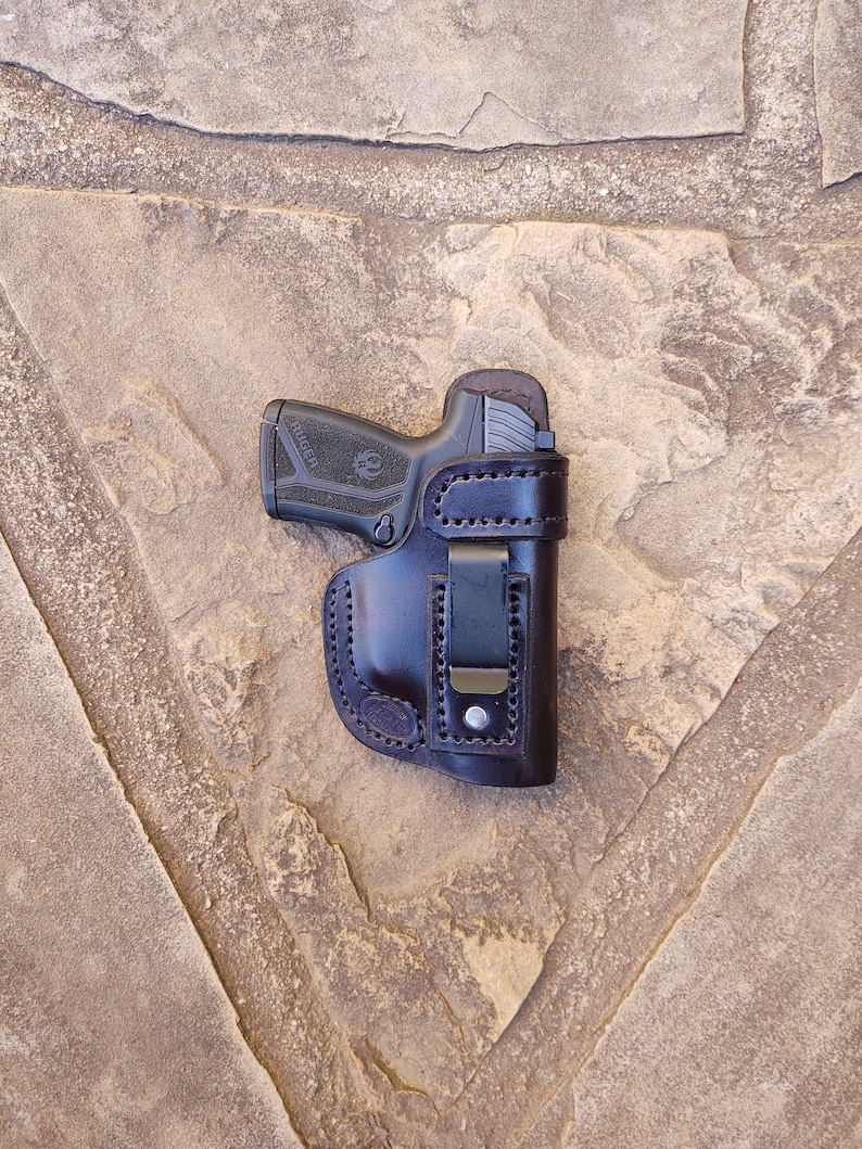 May include: A brown leather holster with a black clip for a handgun. The gun is a black Ruger with a silver trigger.