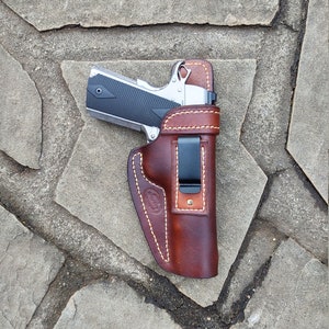May include: A brown leather holster with a stitched edge and a belt clip. A silver handgun is inside the holster.