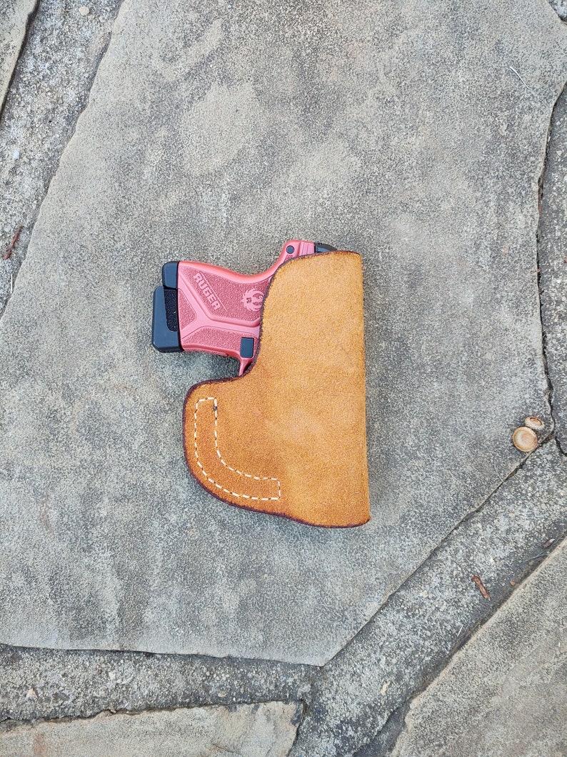 May include: A pink and black handgun in a brown leather holster. The holster has a stitched seam and is designed to be worn on a belt.