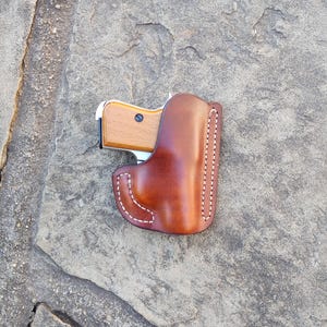 May include: A brown leather holster with a white stitching detail. A small silver and wood handgun is inside the holster.