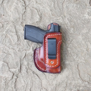 May include: A black handgun in a brown leather holster with white stitching. The holster has a black metal clip. The image is set against a textured, gray stone background.