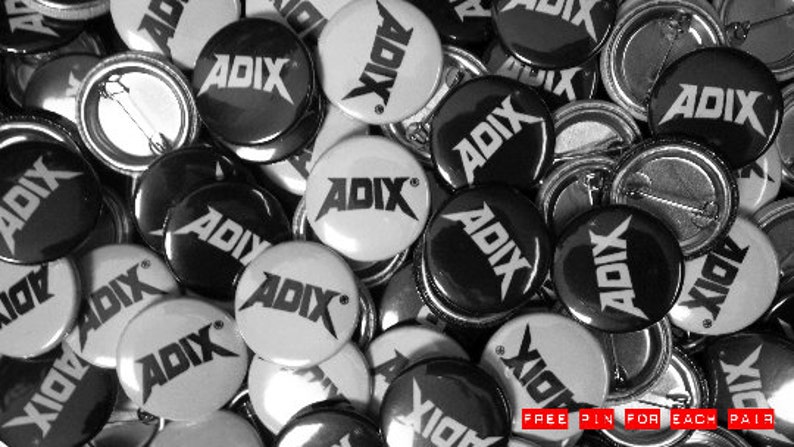 Puede incluir: Una colecci&oacute;n de pines de bot&oacute;n ADIX en blanco y negro. Los pines tienen forma redonda con un alfiler de seguridad en la parte posterior. El logotipo ADIX se muestra en una fuente audaz y estilizada. Tambi&eacute;n se incluye el texto "FREE PIN FOR EACH PAIR".