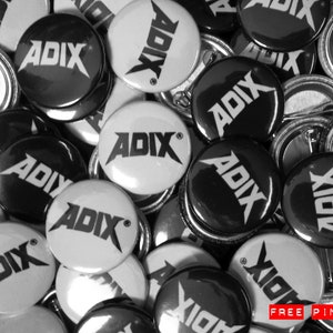 May include: A monochrome image of ADIX logo pin badges. The badges are circular, with a metal pin fastening. The text "ADIX" is displayed in a bold, distinctive font. The image also includes the text "FREE PIN FOR TECH PAIR".