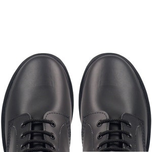 May include: A top-down view of two black leather boots with black laces. The boots have a rounded toe and a textured rubber sole. The boots are set against a white background.