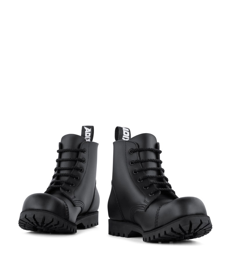 Puede incluir: Un par de botas de combate de cuero negro con suelas gruesas y cordones negros. Las botas tienen una puntera redondeada y una leng&uuml;eta en la parte trasera con la palabra "ADI" impresa. Sobre fondo blanco.