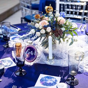 May include: A blue table setting with a centerpiece of white and pink flowers, blue and purple flowers, and greenery. The centerpiece is in a clear glass vase with two white candles. The table is set with white plates, blue napkins, and clear glasses. The table number is "four" and is written on a purple agate slice.