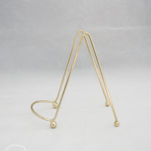 May include: A gold metal display stand with a triangular shape and rounded edges. The stand has three legs and a circular base.