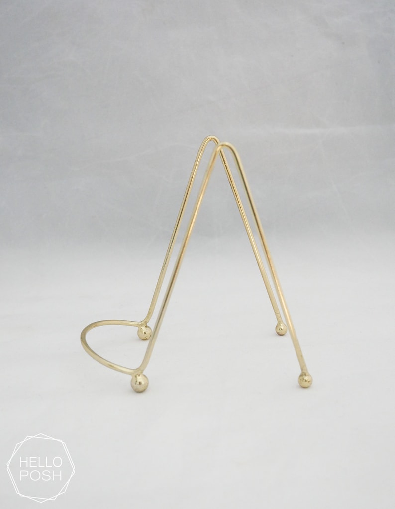 May include: A gold metal display stand with a triangular shape and rounded edges. The stand has three legs and a circular base.