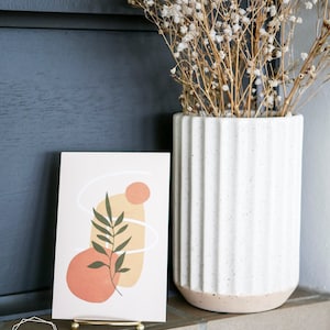May include: A gold metal stand holds a card with an abstract design featuring a green leaf, peach and yellow shapes, and white lines. The card is on a brown surface in front of a white vase with dried white flowers.