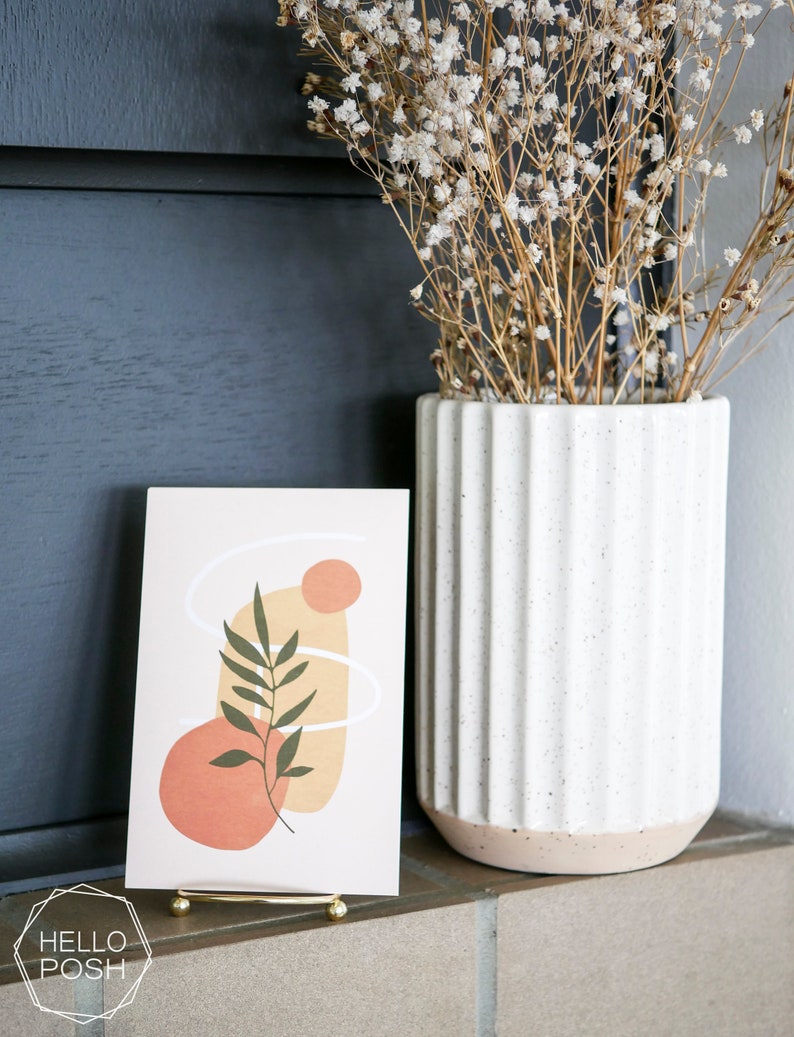May include: A gold metal stand holds a card with an abstract design featuring a green leaf, peach and yellow shapes, and white lines. The card is on a brown surface in front of a white vase with dried white flowers.