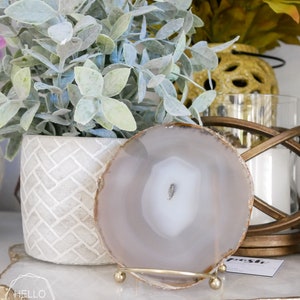May include: A white agate coaster with a gold metal stand. The coaster is on a white surface with a gold and white textured surface.