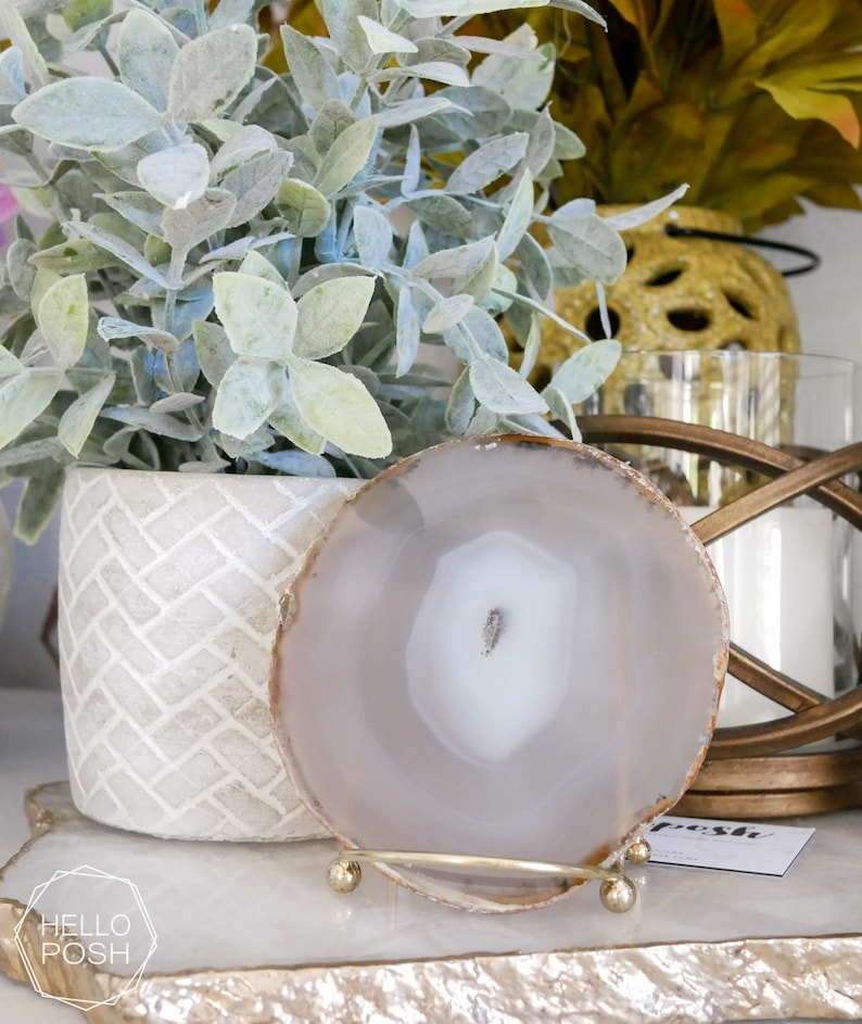 May include: A white agate coaster with a gold metal stand. The coaster is on a white surface with a gold and white textured surface.