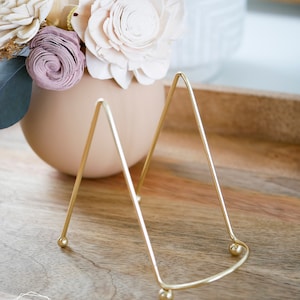 May include: A gold metal display stand with a triangular shape and rounded edges. The stand has three legs and is sitting on a wooden surface.