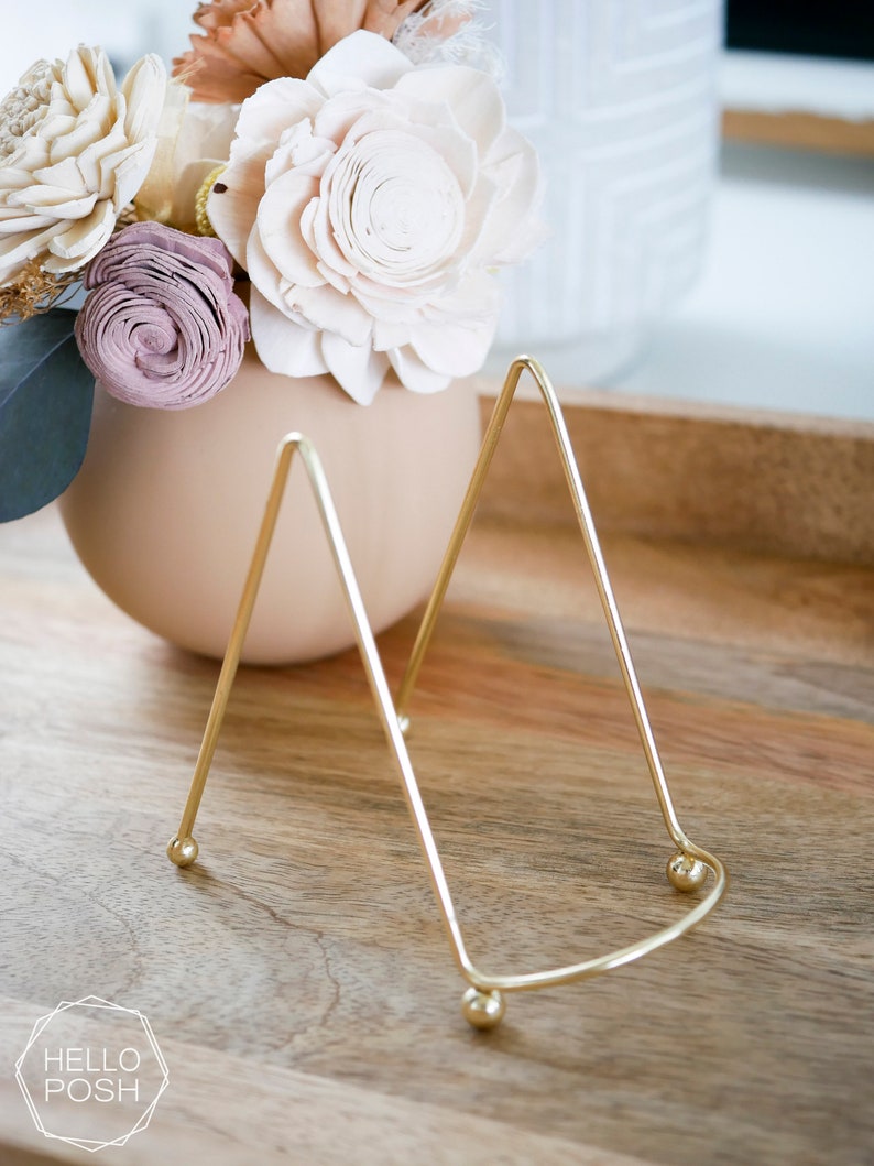 May include: A gold metal display stand with a triangular shape and rounded edges. The stand has three legs and is sitting on a wooden surface.