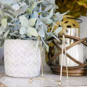 May include: A gold metal picture stand with a triangular base and rounded legs. The stand is on a white marble surface with a gold leaf design. A potted plant with silver-green leaves is in the background.