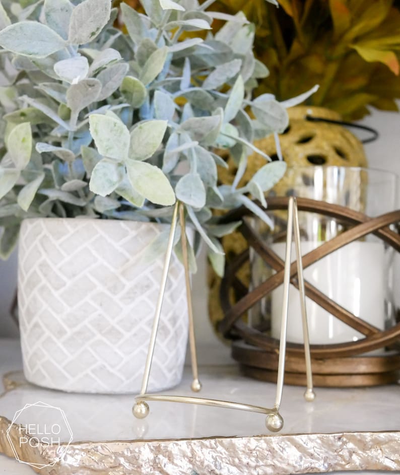 May include: A gold metal picture stand with a triangular base and rounded legs. The stand is on a white marble surface with a gold leaf design. A potted plant with silver-green leaves is in the background.
