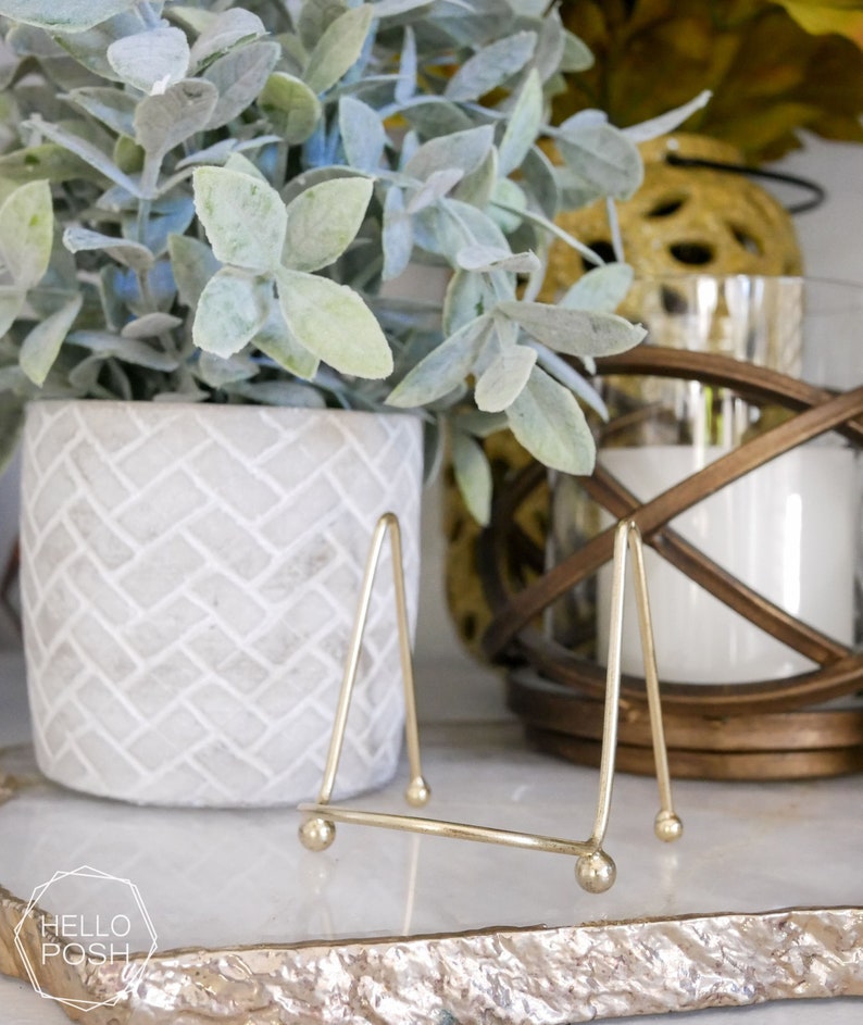 May include: A gold metal picture stand with a triangular shape and rounded feet. The stand is on a white marble surface with a gray concrete planter and a green plant in the background.
