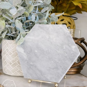 May include: A hexagonal marble coaster with a gray and white veined pattern. The coaster is on a gold metal stand with three legs.