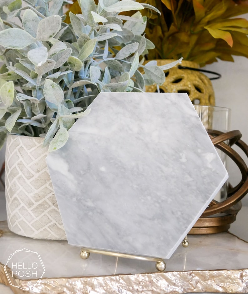 May include: A hexagonal marble coaster with a gray and white veined pattern. The coaster is on a gold metal stand with three legs.