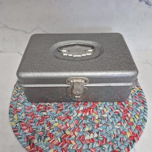 May include: A rectangular, silver-colored metal box with a handle and clasp closure. The box sits on a round, braided trivet with a pattern of red, blue, yellow, and white. The box has a textured surface.
