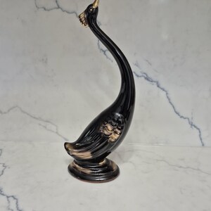 May include: A glossy black ceramic figurine of a swan with an elongated, curved neck. The swan features gold-toned accents on its beak, eye, and wing feathers, and it stands on a circular base. The sculpture has a smooth, reflective finish.