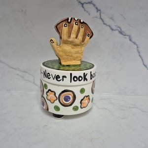 May include: A ceramic trinket box with a hand-shaped lid holding a football. The box is white with colorful floral designs and the words "Never look ba" written on the side. The hand is tan and the football is brown.