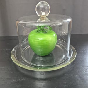 May include: A bright green glass apple figurine with a clear glass stem and leaf detail, enclosed within a clear glass cloche dome that features a spherical handle on top, resting on a matching circular glass plate base.