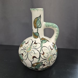 May include: A cream-colored ceramic pitcher with a rounded body and a single handle. The surface is decorated with hand-painted white dogwood flowers featuring brown outlines, green leaves, and a speckled green texture on the handle and rim.