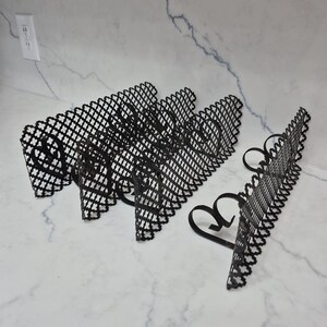 May include: Black metal decorative plate holders with a heart-shaped base and a decorative, openwork design. The holders are arranged on a white marble surface. The holders are designed to display plates.