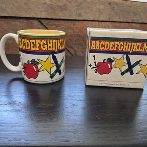 May include: A white ceramic mug and a matching square box, both featuring a yellow interior rim, a red band with the alphabet in yellow, and a graphic pattern of a red apple, yellow stars, and a blue crayon on a white background.