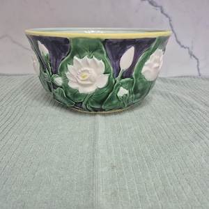 May include: A ceramic bowl with a floral design. The bowl features white lotus flowers and green leaves against a dark blue background. The rim is yellow. The bowl is likely used for serving or display.