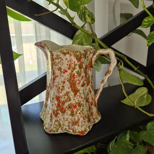 May include: A vintage ceramic pitcher with a cream base and a speckled pattern of red and green. The pitcher has a scalloped rim and a curved handle. It is displayed on a black shelf with green plants.