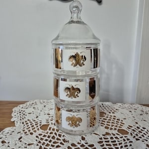 May include: A clear glass three-tiered stackable canister set with a matching lid. Each of the three cylindrical sections features a white rectangular panel decorated with a gold-colored fleur-de-lis emblem and gold-colored vertical side accents.
