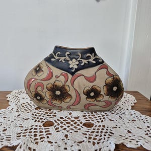 May include: A decorative ceramic vase with a cream-colored base, adorned with brown floral designs and pink ribbon accents. The top features a dark blue band with white floral patterns. The vase is displayed on a wooden surface.