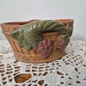 May include: A round ceramic bowl with a textured basket-weave pattern in a light brown finish. The exterior features a decorative relief of green leaves and clusters of purple grapes wrapped around the upper rim of the vessel.