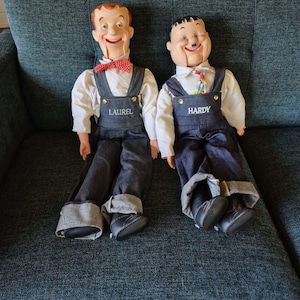 May include: Two ventriloquist-style dolls representing Laurel and Hardy. Each doll wears a white long-sleeved shirt, dark denim overalls with gold buttons, and black shoes. The left doll has a red polka-dot bow tie and the text LAUREL on the bib.