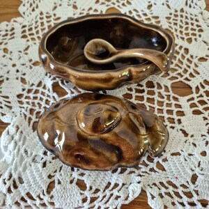 May include: A brown glazed ceramic lidded bowl with an irregular, organic shape and a matching small spoon resting inside. The bowl features a glossy, mottled finish with darker and lighter brown tones, and a lid with a central knob handle.