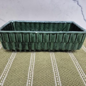 May include: A rectangular, dark green ceramic planter with a textured, woven-like pattern. The planter has a slightly glossy finish and is set on a green and white striped surface. Ideal for small plants or succulents.