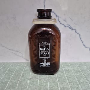 May include: A brown glass bottle with a dark brown cap and a white label. The label reads "The Gates Homestead Farms 1798". The bottle has a rectangular shape with rounded edges and a narrow neck. The bottle is sitting on a green surface.