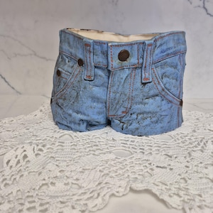 May include: A ceramic planter shaped like a pair of blue denim shorts. The planter has brown buttons, pockets, and orange stitching details. The denim has a distressed look, and the planter sits on a white lace doily.