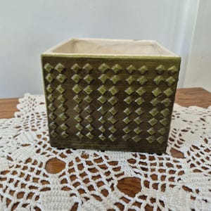 May include: A square, olive green ceramic planter with a repeating diamond pattern. The interior is a cream color. The planter sits on a white, crocheted doily. The planter is suitable for small plants or succulents.